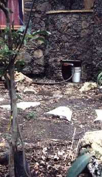 Gray water tank incorporated in outside wall of house, showing gray water hose and bucket below.